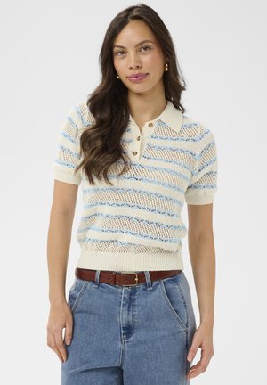 Jonge vrouw met lang haar die een gebreide polo met crèmekleurige en lichtblauwe strepen draagt, een bruine riem en een high-waisted blauwe spijkerbroek, staand tegen een witte achtergrond.