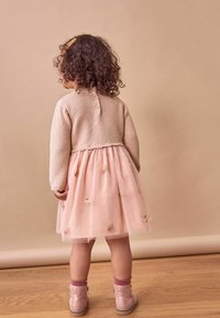 Helles rosa Kleid mit Tüllrock und floralen Stickereien, kombiniert mit einer beige gestrickten Strickjacke. Rosa Knöchelstiefel mit Reißverschlüssen und passenden Socken.