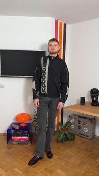 Μαύρο μπουφάν με φερμουάρ και γκρι πάνελ με λεπτομέρεια λογότυπου στη μία πλευρά, συνδυασμένο με γκρι τζιν καμπάνα και σκούρα παπούτσια. Απλά στοιχεία φόντου.