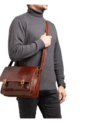 Uomo che indossa un dolcevita grigio e pantaloni neri, con una borsa a tracolla in pelle marrone con fibbie in ottone.