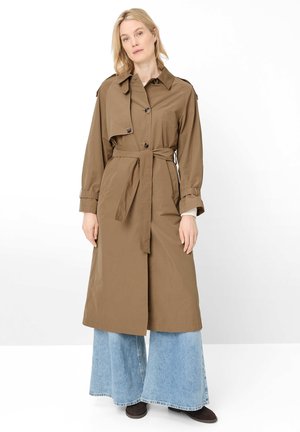Blonde woman wearing a brown belted trench coat over light blue wide-leg jeans and dark shoes, standing against a white background.