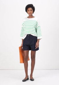 Oversized Pullover in Weiß und Grün gestreift, marineblaue, gefaltete Shorts, schwarze Ballerinas und orangefarbene Tote-Bag. Klare Linien und kontrastierende Farben.