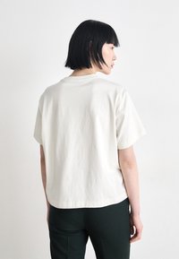 Cremefarvet bomulds t-shirt med korte ærmer, afslappet pasform og rund halsudskæring, båret med mørkegrønne bukser. Glat tekstur og ensartet design.