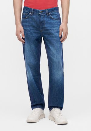 Homme portant un jean droit bleu, des baskets blanches et une chemise rouge, debout devant un fond clair uni.
