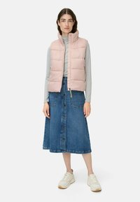 Rosa gepolsterte Weste mit hohem Kragen über einem grauen Langarmshirt, kombiniert mit einem Denimrock mit Knopfleiste und weißen klobigen Sneakers.