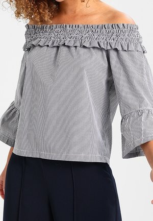 Frau trägt eine schwarz-weiß karierte Bluse mit ausgeschnittenen Schultern, gerafftem Ausschnitt und dreiviertelärmel, kombiniert mit dunkler Hose.