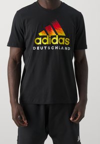 Czarny bawełniany T-shirt z gradientowym logo Adidasa w kolorach żółtym, czerwonym i pomarańczowym oraz napisem "DEUTSCHLAND" poniżej, krótki rękaw, standardowy krój.