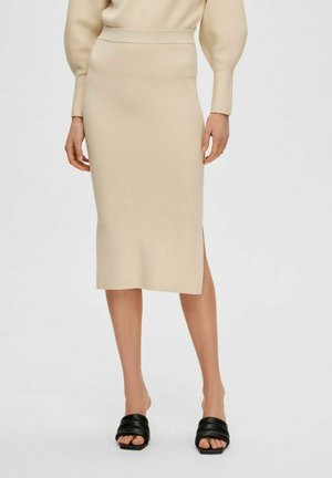 Beige pletená midi sukně s přiléhavým pasem a bočním rozparkem, kombinovaná s černými skluznými sandály. Hladká textura, přizpůsobený design.
