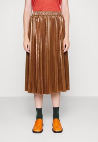 Bronsskörta med veck och en skimrande textur, tillsammans med orange skor och gröna strumpor. Blusen är röd och instoppad i midjan.