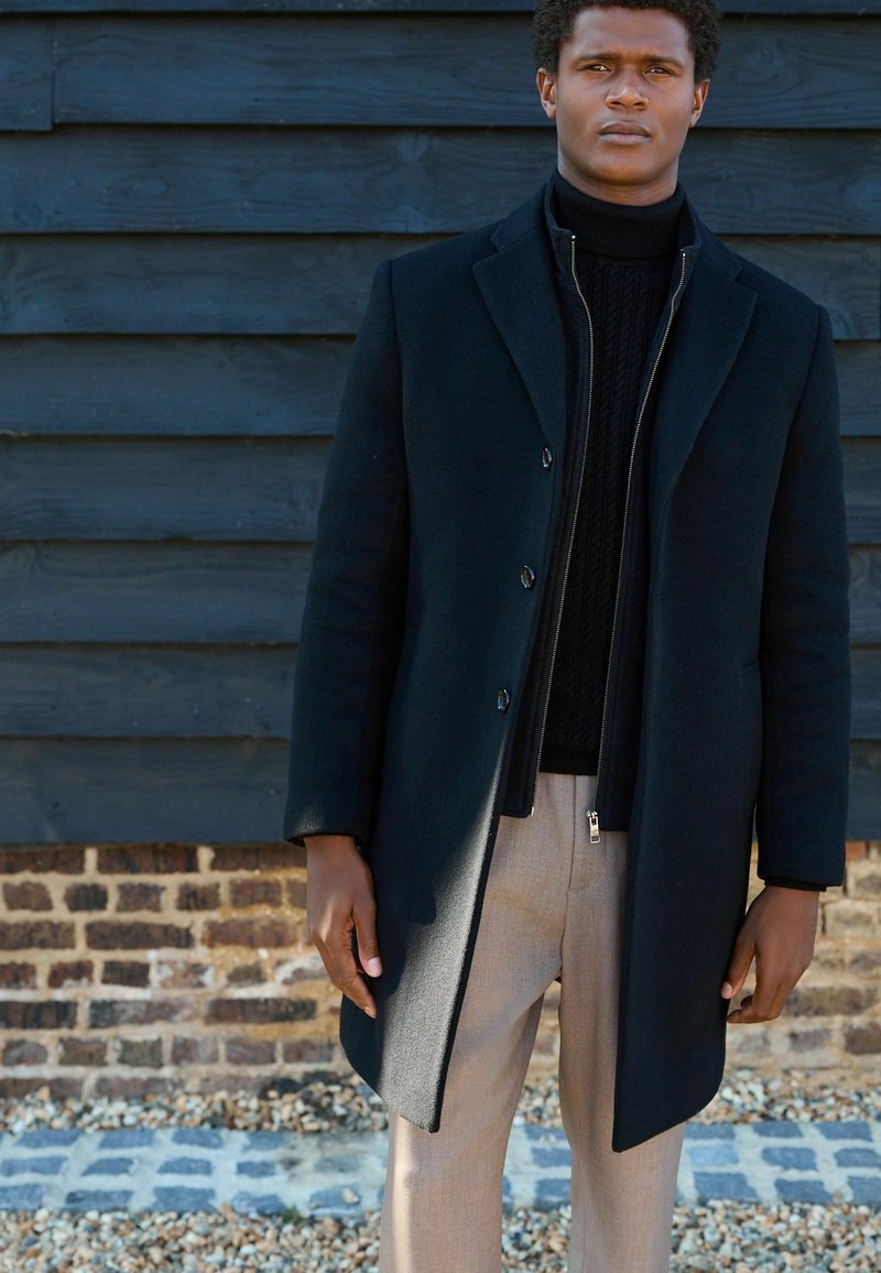 Langer schwarzer Mantel mit maßgeschneidertem Design, über einem schwarzen Strickpullover getragen. Beigefarbene Hose vervollständigt den Look. Glatte Textur und minimalistische Details.