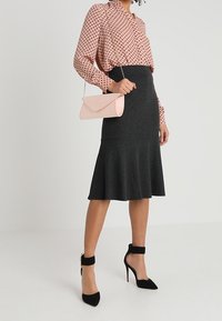 Femme portant un chemisier rose à pois, une jupe midi évasée gris foncé, des talons noirs à bride cheville, et tenant une petite pochette rose.