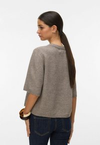 Femme avec une longue queue de cheval noire portant un pull gris à manches courtes en tricot, un jean bleu foncé et de grands bracelets au poignet, tournée vers la gauche.