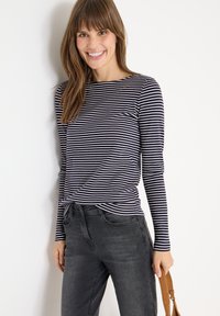 Granatowa i biała w pionowe paski bluzka z długim rękawem oraz okrągłym dekoltem, z wiązanym detalem w talii, zestawiona z ciemnoszarymi dżinsami i jasnobrązową torebką.