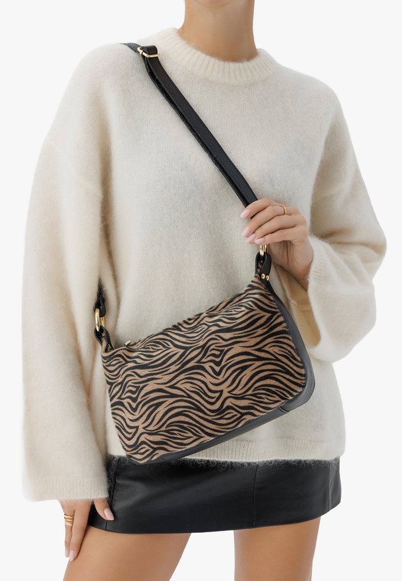 Borsa a spalla con stampa zebra marrone e nera, realizzata in un tessuto testurizzato, con tracolla nera e finiture in metallo dorato, abbinata a un maglione crema e una gonna nera.
