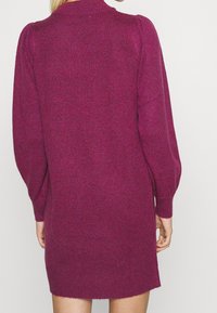 Robe pull tricotée en magenta avec une finition texturée, présentant un col haut, de longues manches bouffantes et un ourlet légèrement effiloché.
