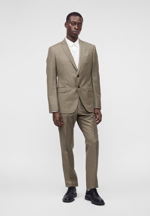 Homme debout portant un costume beige, une chemise blanche et des chaussures noires sur un fond clair uni.