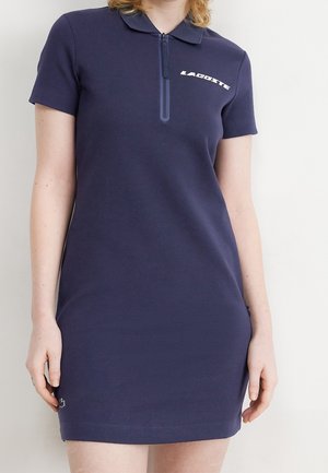 Mujer vistiendo un vestido tipo polo azul marino de manga corta con cremallera frontal y logo blanco "Lacoste" en el pecho.