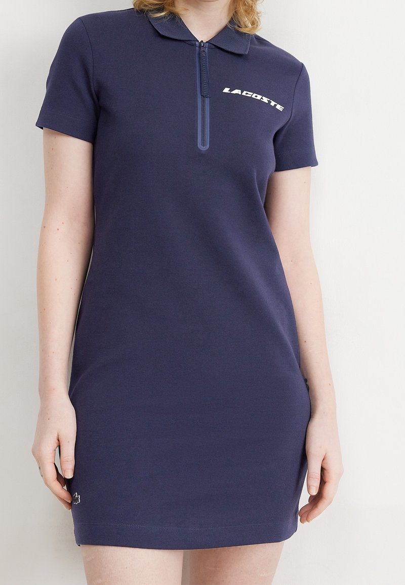 Femme portant une robe polo bleu marine à manches courtes avec une fermeture éclair devant et un logo blanc "Lacoste" sur la poitrine.