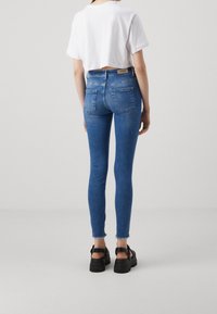 Jeans skinny de color azul con un dobladillo deshilachado, cintura alta y dos bolsillos traseros. Combinados con una camiseta blanca recortada y sandalias negras gruesas.