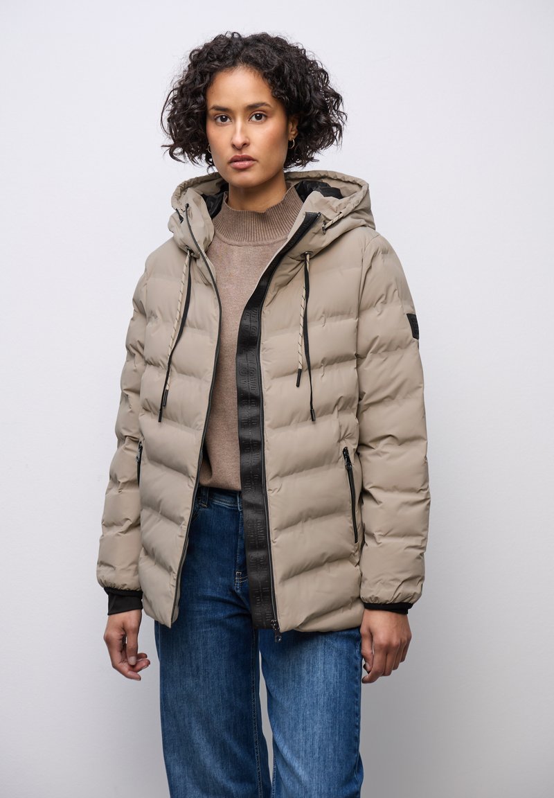 Beige dynejakke med sort lynlås, med hætte og elastiske manchetter. Den har et quiltet mønster og sidelommer. Bruges over en brun sweater.