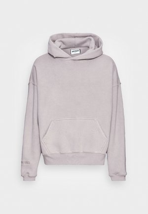 Personne portant un sweat à capuche beige et un jean bleu avec un motif d'aigle, debout sur le bord du trottoir dans une zone urbaine avec des murs tagués.