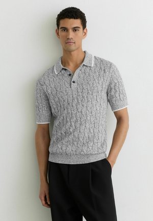 Noor mees lühikeste varrukatega halli kaabel-kootud kraega polo särgis valgete äärtega, mustade pükstega, seisab valge tavalise seina taustal.