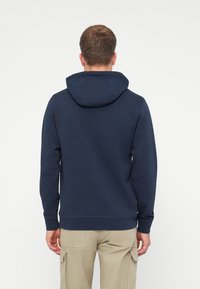 Μπλούζα ναυτικού με επενδυμένη κουκούλα, ριμπ μανσέτες και στρίφωμα. Μαλακό ύφασμα με λείο τελείωμα, φορεμένο πάνω από ανοιχτό καφέ cargo παντελόνι.