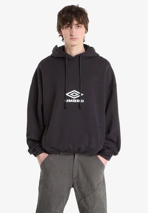 Giovane uomo in piedi con le mani nelle tasche, indossa una felpa nera Umbro con cappuccio e pantaloni grigi su sfondo bianco uniforme.