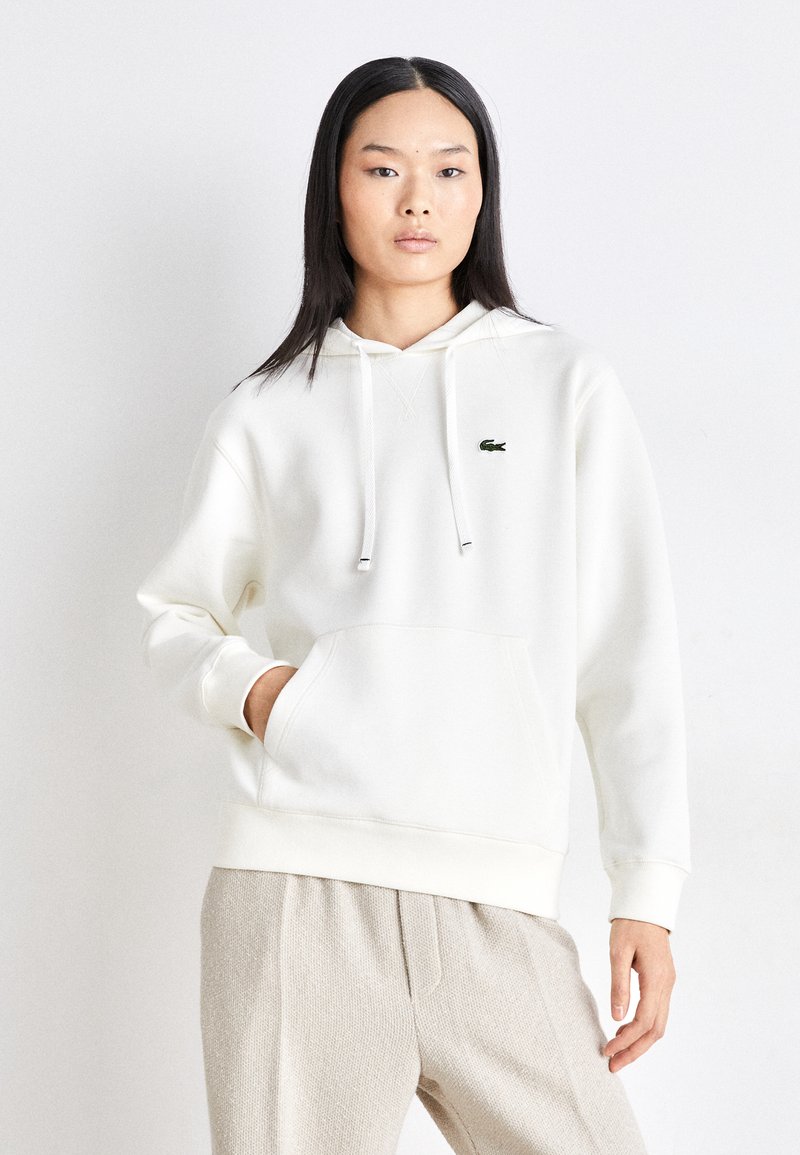 Felpa bianca con tasca frontale e coulisse, caratterizzata da un piccolo logo verde Lacoste. Abbinata a pantaloni beige chiaro con texture.