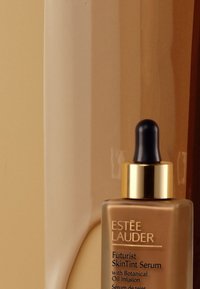 Brun glassflaske med Estée Lauder Futurist SkinTint Serum med pipette. Gullfargede detaljer på en beige bakgrunn med tekst på etiketten.