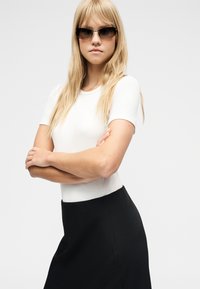 Νεαρή γυναίκα με μακριά ξανθά μαλλιά, που φορά σκούρα γυαλιά ηλίου, λευκό μπλουζάκι με κοντά μανίκια και μαύρη φούστα, στεκόμενη με σταυρωμένα τα χέρια.