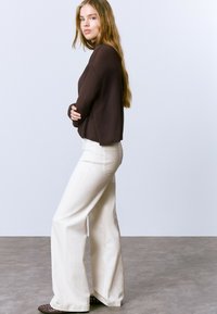 Camiseta de punto de manga larga marrón combinada con pantalones de pierna ancha blancos y zapatos oscuros. La modelo está de pie con los brazos cruzados contra un fondo liso.