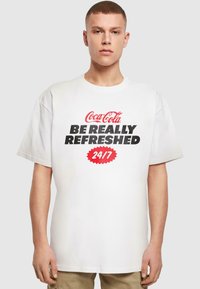 Hvid bomulds T-shirt med en iøjnefaldende sort og rød Coca-Cola grafik. Teksten lyder "BLIV RIGTIG FORNYET 24/7". Klassisk rund halsudskæring og afslappet pasform.