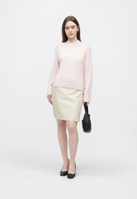 Mladá žena v světle růžovém svetru, béžové sukni a černých lodičkách, držící malou černou kabelku, stojí před jednobarevným bílým pozadím.