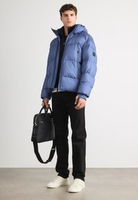 Giacca a vento blu con cappuccio, tasche con zip e polsini a costine. Pantaloni neri e sneakers di colore chiaro. Tieni in mano una borsa nera con tracolla.