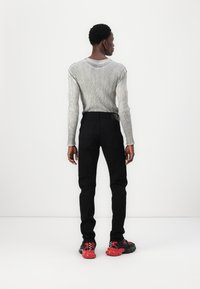 Muž stojaci chrbtom, oblečený v rebrovom šedom tričku s dlhým rukávom, čiernych nohaviciach a čiernych teniskách s červenými podrážkami.