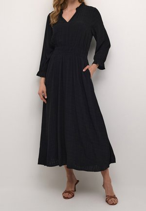 Kobieta ubrana w długą czarną sukienkę z dekoltem w serek, marszczoną talią i mankietami, stojąca z jedną ręką w kieszeni, w połączeniu z brązowymi sandałami na obcasie.