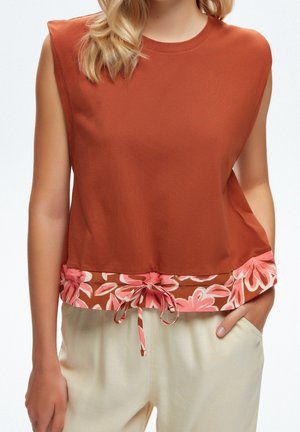 Ärmelloses rostfarbenes Top mit rosa floralem Saum und Kordelzug, getragen mit hellbeigen Hosen, Hand in der Tasche, aufrechte Haltung.