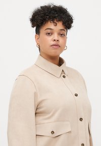 Beige suede shirt jacket with a collar, front button closure, and two patch pockets. Features stitched detailing and round buttons.