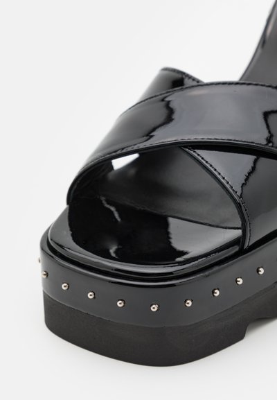 Sandalias de plataforma negras con acabado de charol brillante, tiras entrelazadas y tachuelas plateadas a lo largo de la suela para un detalle adicional.