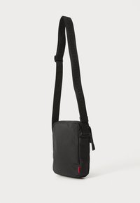 Borsa a tracolla nera realizzata in tessuto testurizzato, con chiusura a zip e spallaccio con marchio. Accento rosso nell'angolo in basso a sinistra. Forma compatta e rettangolare.