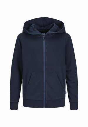 Barn iført mørkeblå hættetrøje med lynlås, hvid T-shirt med grafik, løstsiddende mørke bukser og hvide sneakers med grøn stribe, stående mod en ensfarvet baggrund.