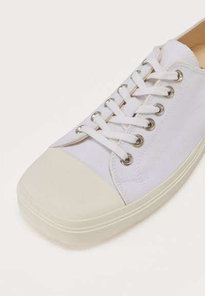 Valge kanvas sneaker ümmarguse beeži kummi varbaga, hõbedaste silmade ja valgete paeltega. Omab siledat tekstuuri ja madalat profiili.