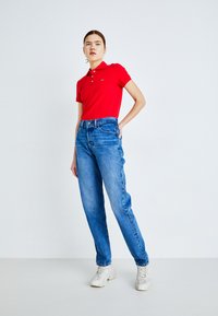 Camisa polo roja con cuello, combinada con jeans de corte holgado en azul claro y zapatillas grises. Atuendo casual sobre un fondo simple.