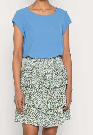 Vrouw die een lichtblauw T-shirt met korte mouwen en een gelaagde rok met patronen in groene en lichtblauwe tinten draagt.