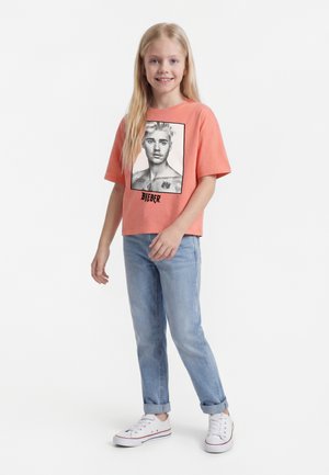 Fiatal szőke lány korall színű pólóban, amelyen fekete-fehér portré van, és világoskék farmernadrágban, mosolyogva, egyik kezét a derekánál tartva áll.