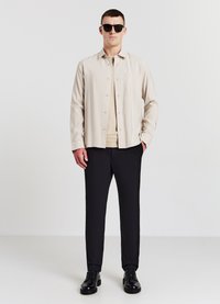 Beige knopfnahe Hemd, schwarze Hose, schwarze Schuhe. Sonnenbrille und eine Tasche auf dem Hemd. Glatte Stoffstruktur, maßgeschneiderte Passform.