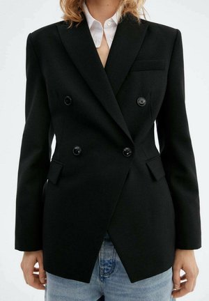 Femme portant un blazer noir cintré à double boutonnage sur une chemise blanche et un jean bleu clair, debout devant un fond uni.