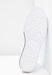 Semelle de basket blanche avec un motif de bande de roulement géométrique texturé et une bande noire le long du bord, positionnée verticalement contre un coin blanc.