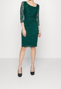 Femme vêtue d'une robe en dentelle vert foncé longueur genou avec des manches trois-quarts et des chaussures à talons noirs, debout devant un fond uni.